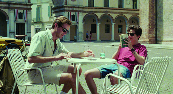 Two men sit at a white metal cafe table in a sunny outdoor plaza with arched buildings in the background.