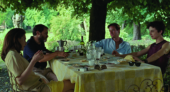 Four people sit around a yellow checkered table for an outdoor meal under a large leafy tree.