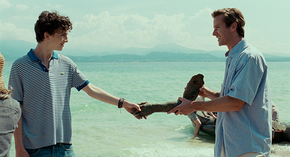Two men stand on a beach by turquoise water, one handing a large piece of driftwood to the other.