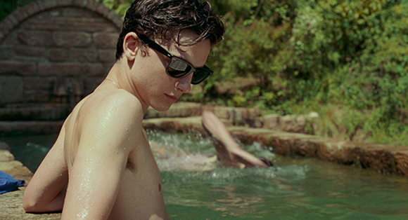 A shirtless young man in sunglasses sits by the edge of a stone pool with greenery in the background.