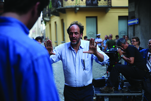 A man in a light blue shirt stands with both hands raised in a gesturing motion on a film set.