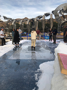 A group of people stand on a large, melting ice platform surrounded by snow and large metal structures.