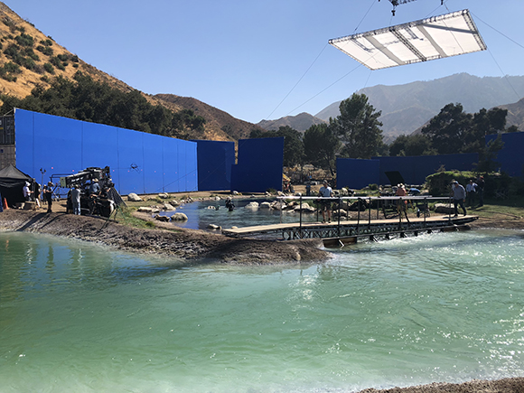 Film crew working on a set with a blue screen, a small pond, and mountains in the background.