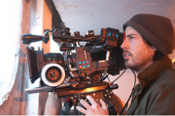 A man in a brown beanie looks through the viewfinder of a large professional cinema camera.