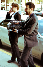 Two men in dark suits stand by a white car; one holds a professional video camera while the other talks on a phone.