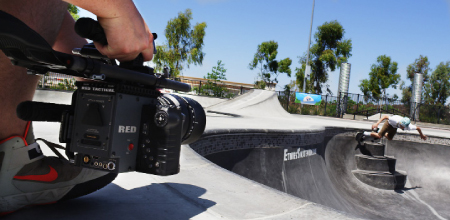 A person holds a professional RED cinema camera, filming a skateboarder performing a trick in a concrete bowl.