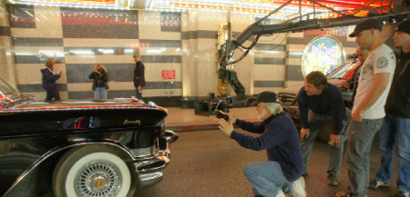 Film crew uses a camera crane to film a black vintage car in a large, tiled lobby.
