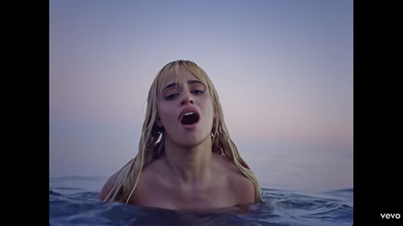 A blonde woman with wet hair emerges from the ocean water against a twilight sky.
