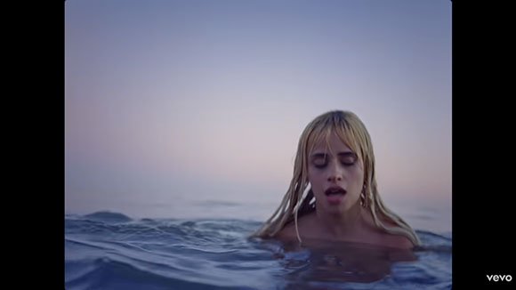 A woman with wet blonde hair rises from dark ocean waves under a twilight sky.