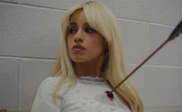 A blonde woman in a white lace top looks away as an arrow is lodged in her shoulder.