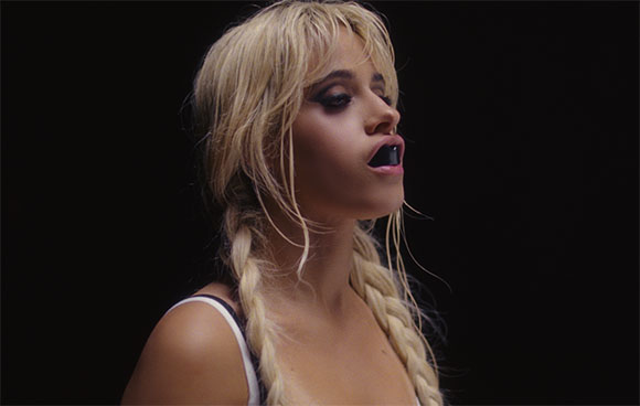 A woman with blonde hair in two braids and dark eye makeup looking to the side against a black background.