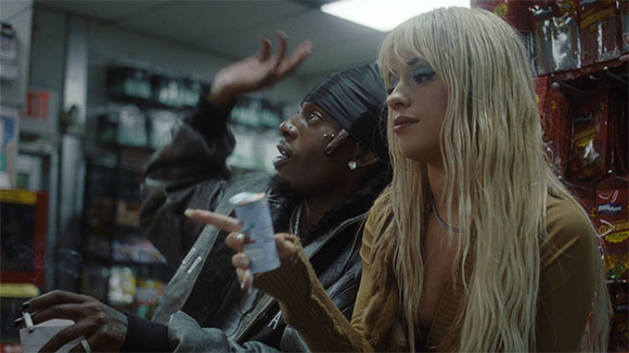 A man in a black headwrap gestures while a blonde woman holds a small container in a store.