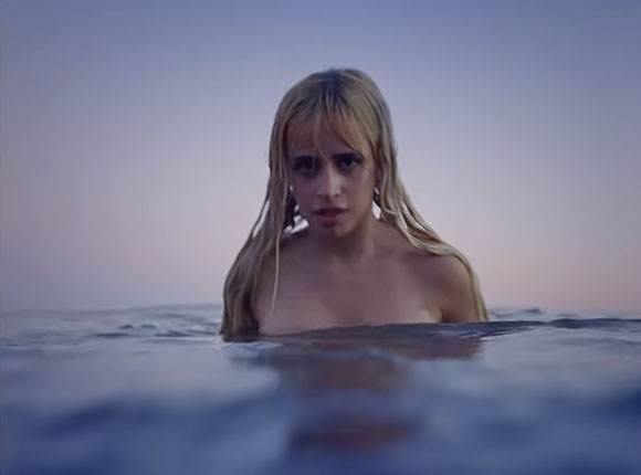 A blonde woman looks directly at the camera while waist-deep in calm water under a twilight sky.