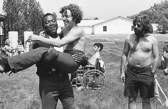 A man carries another man in his arms in a grassy field with people and a wheelchair in the background.