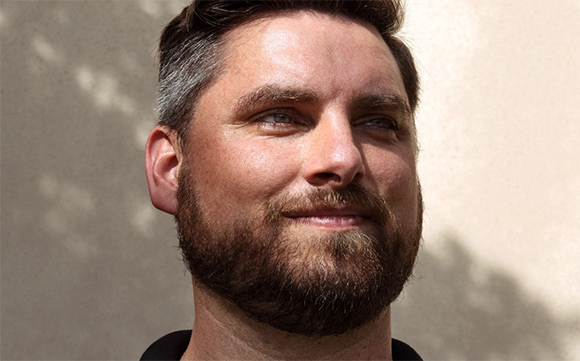 A close-up portrait of a man with short dark hair and a brown beard looking slightly to the side.