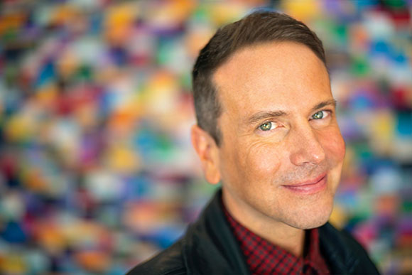 A man with short dark hair and green eyes smiles toward the camera against a blurred, multicolored background.