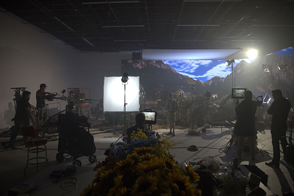 Film crew members working in a studio with large lighting equipment and a mountain landscape backdrop.