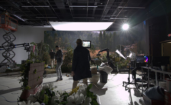 Film crew members working on a studio set featuring a large forest landscape backdrop and studio lighting.