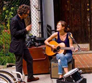A man in a black jacket talks to a woman sitting with an acoustic guitar and amplifier.