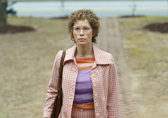 A woman with curly hair and large glasses wears a pink checkered jacket over a striped shirt outdoors.