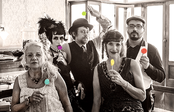 Five people in vintage costumes holding colorful lollipops in a sepia-toned indoor setting.