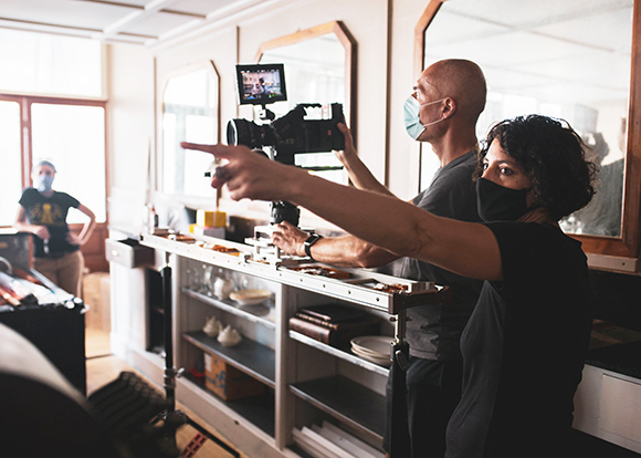 Two crew members wearing masks operate a professional camera on a tripod in a bright studio.