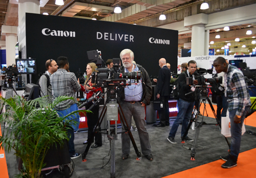 A film crew with cameras and tripods works in a convention hall in front of a large black Canon sign.