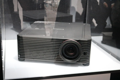 A dark gray Canon projector with a large lens is displayed on a white surface, enclosed by a clear barrier. The Canon logo is