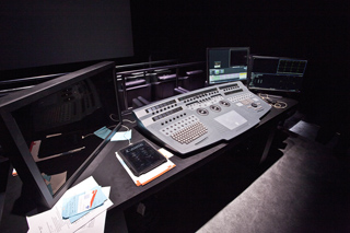 A large grey video editing console sits on a black desk with three computer monitors in a dark studio.