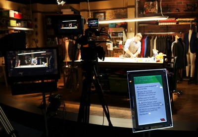 A professional video camera on a tripod is in the foreground, with a monitor showing a person at a table and a tablet display
