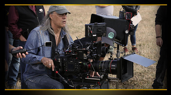Cinematographer with long gray hair and a denim shirt operates a complex camera rig with multiple lenses and monitors.