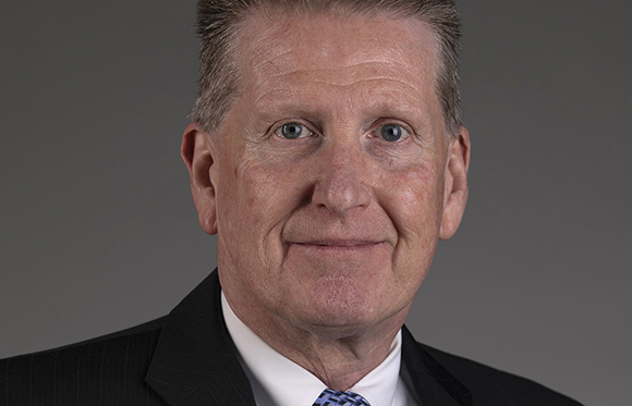 A man with light skin and short brown hair wearing a black suit and white shirt.