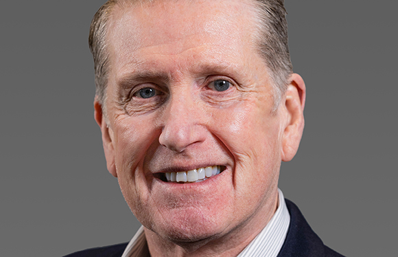 A smiling man with light hair and blue eyes looking toward the camera against a gray background.