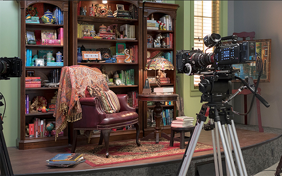 A professional film camera on a tripod overlooks a library-themed studio set with a leather chair and bookshelves.