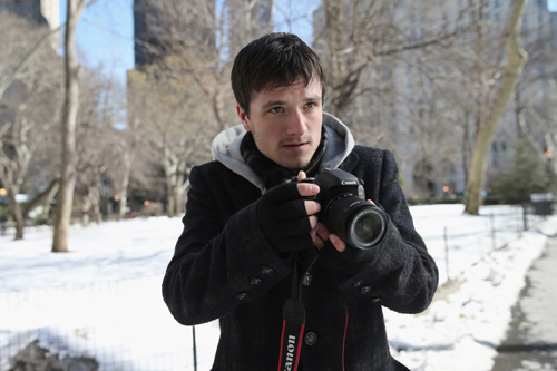 A man in a dark coat and gloves holds a Canon DSLR camera in a snowy outdoor park setting.