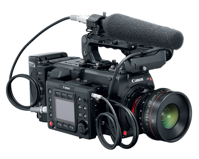 A black Canon professional video camera equipped with a large lens and a shotgun microphone with a foam windscreen.