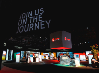 A large Canon exhibition booth under a dark sky with 'JOIN US ON THE JOURNEY' projected in large white letters.