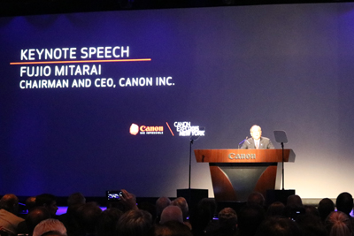 A man stands at a wooden podium with the Canon logo, delivering a keynote speech. The screen behind him displays "KEYNOTE SPE