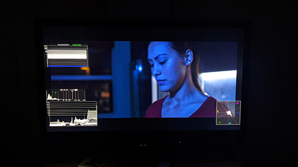 A dark room shows a monitor displaying a woman's face alongside audio waveforms and color grading tools.