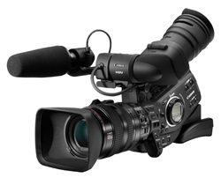 Professional black video camera equipped with a large lens and a shotgun microphone on top.