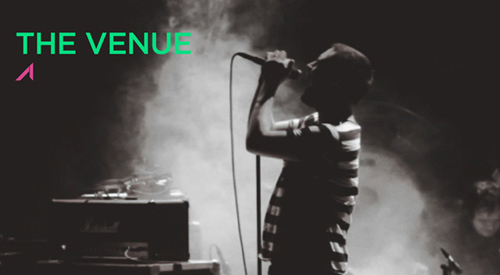 A person in a striped shirt sings into a microphone on a stage with smoke in the background.