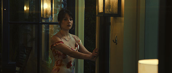 A woman in a floral dress stands in a doorway, looking out into the darkness at night.