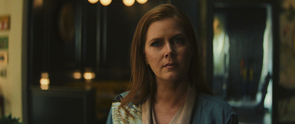 A woman with long red hair looks forward with a neutral, serious expression in a dimly lit room.