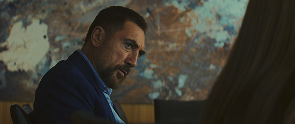 A man with a beard and dark hair wearing a blue suit looks intently toward a person in the foreground.