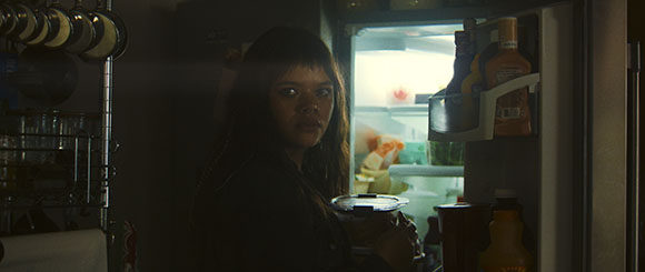 A woman with dark hair looks toward the camera in a dark kitchen next to an open refrigerator.