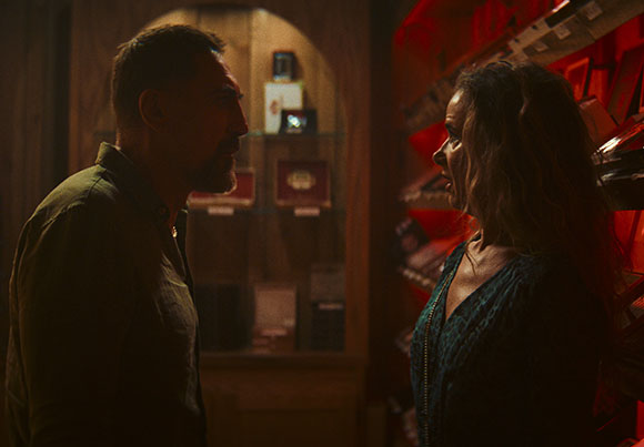 A man and a woman stand close together, looking at each other in a room with warm lighting.