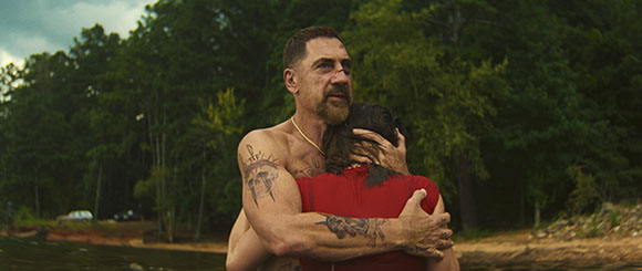 A shirtless, tattooed man with a beard embraces a woman in a red shirt by a lake with trees in the background.