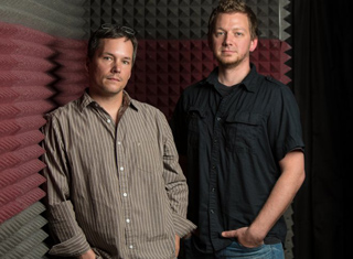Two men standing side by side in front of acoustic foam padding in a studio.