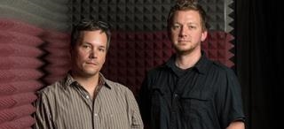 Two men standing side by side in a room with acoustic foam padding on the walls.