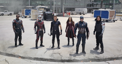 Six costumed superheroes stand in a circle on a concrete lot with vehicles and equipment in the background.